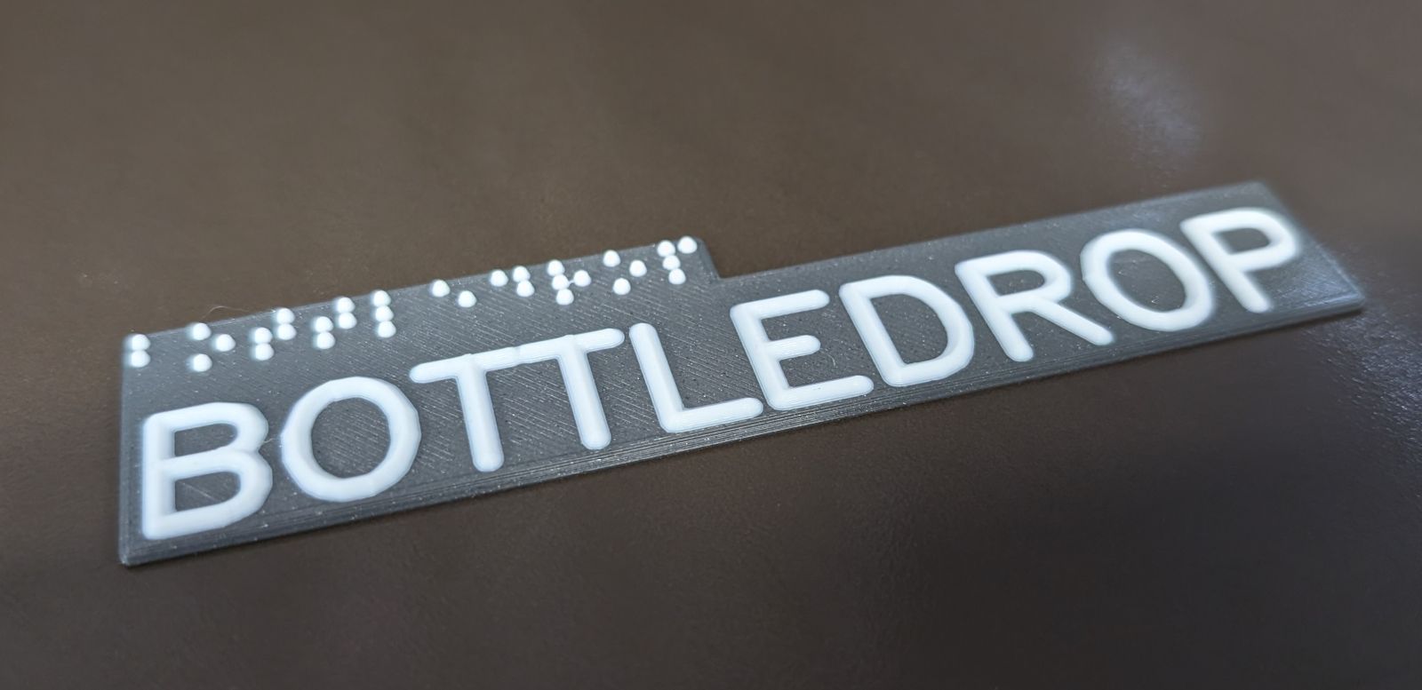 Ein Schild
		mit dem Text Bottledrop in Profilschrift und Braille liegt auf dem Bett eines
		3D-Druckers. Es hat einen grauen Hintergrund und die Schrift ist weiß